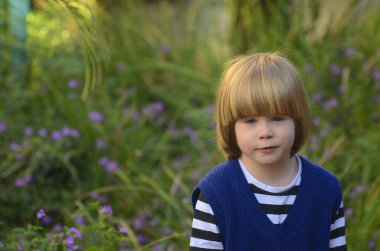 Sarı uzun saçlı, gülümseyen, gülümseyen, çekici bir çocuğun portresi. Arka planda çimenler ve mor çiçekler var. Anneler günü kartı, bahar günü kartı, anaokulu reklamı.
