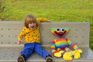 A positive little boy, 4 years old, walks in the park with a bright colored toy. Concept: kindergarten, imaginary friend, favorite toy. The child sits on a bench with a very large soft toy.