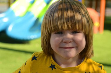 Front view of small caucasian boy four years on playground. The boy smiles and laughs. Portrait of a child on a walk. A boy in a yellow sweater on the background of a slide