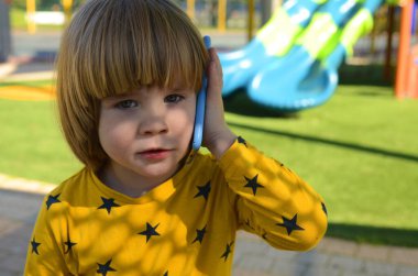A little boy calls on a smartphone. Child on the playground, slide in the background. boy calling parents, talking with friends