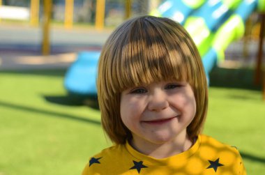 Front view of small caucasian boy four years on playground. The boy smiles and laughs. Portrait of a child on a walk. A boy in a yellow sweater on the background of a slide