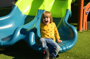 Front view of small caucasian boy four years on playground. The boy smiles and laughs. Portrait of a child on a walk. A boy in a yellow sweater on the background of a slide
