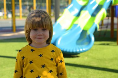 Front view of small caucasian boy four years on playground. The boy smiles and laughs. Portrait of a child on a walk. A boy in a yellow sweater on the background of a slide
