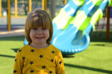 Front view of small caucasian boy four years on playground. The boy smiles and laughs. Portrait of a child on a walk. A boy in a yellow sweater on the background of a slide