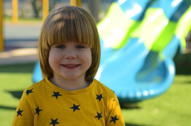 Front view of small caucasian boy four years on playground. The boy smiles and laughs. Portrait of a child on a walk. A boy in a yellow sweater on the background of a slide