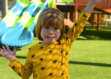 Front view of small caucasian boy four years on playground. The boy smiles and laughs. Portrait of a child on a walk. A boy in a yellow sweater on the background of a slide