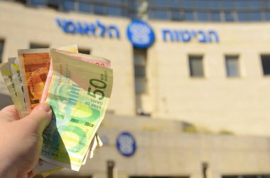 Logo Bituah Leumi. The hand holds money, new Israeli shekel. payments to Bituach Leumi. Israeli national social services. Translate: National Insurance Institute . Holon Israel February 2023
