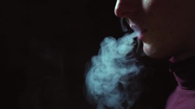 Sigara içmek için elektrikli cihaz. Karanlıkta bir adam elektronik sigara içiyor. Vape. E-sigara, vsping. Kavram: Sigara içmenin zararı. Yavaş çekim, karanlık arka planda sigara, yüzü olmayan bir adam elinde elektronik bir sigara tutuyor.