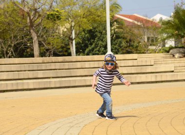Çizgili tişörtlü ve güneş gözlüklü havalı bir çocuk. 4 yaşında bir çocuk parkta koşup dans ediyor. Aktif pozitif çocuk. Keyfin yerinde yaz..