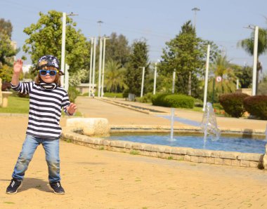 Çizgili tişörtlü ve güneş gözlüklü havalı bir çocuk. 4 yaşında bir çocuk parkta koşup dans ediyor. Aktif pozitif çocuk. Keyfin yerinde yaz..