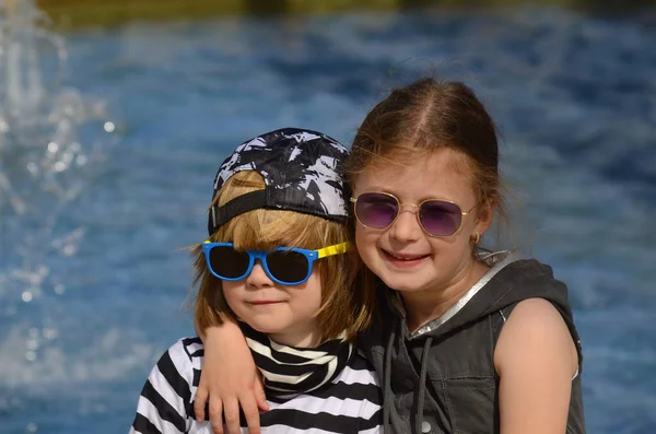 孩子们在游泳池 喷泉的后面 戴着太阳镜的小男孩和小女孩 女孩拥抱着男孩 兄弟和姐妹 度假的朋友 探亲旅行 — 图库照片
