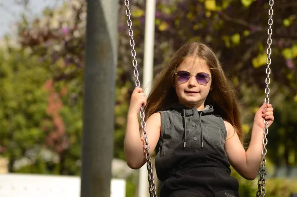 stock image A girl with blond long hair in sunglasses rides on a swing in the spring. Girl 6 years old. Portrait of a child on the playground in the garden