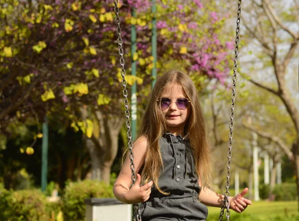 一个戴着太阳镜的金发女孩骑在秋千上 6岁的女孩花园里操场上一个孩子的画像 — 图库照片