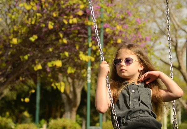 一个戴着太阳镜的金发女孩骑在秋千上 6岁的女孩花园里操场上一个孩子的画像 — 图库照片