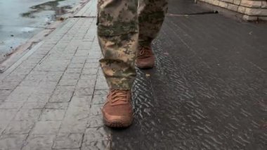 Ukraynalı asker yağmurda sokakta yürüyor. Askeri botlar, sırt çantası, Ukrayna bayrağı. Silahlı kuvvetler, vatanın savunucusu.