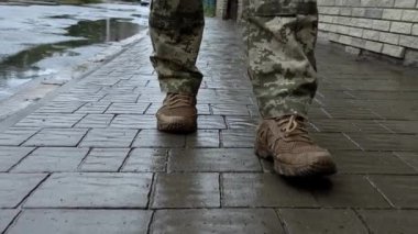 Ukraynalı asker yağmurda sokakta yürüyor. Askeri botlar, sırt çantası, Ukrayna bayrağı. Silahlı kuvvetler, vatanın savunucusu.