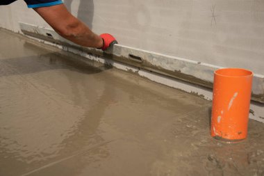 Mason leveling and screeding concrete floor base with square trowel in front of the house. Construction business, do-it-yourself concept. Hand in red glove. clipart