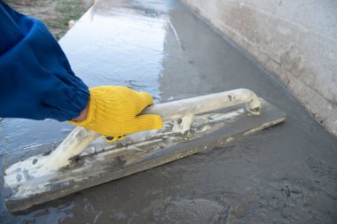 Mason evin önündeki kare malayla beton zemini düzleştiriyor ve tarıyor. İnşaat işi, kendin yap konsepti. Sarı eldivenli el..