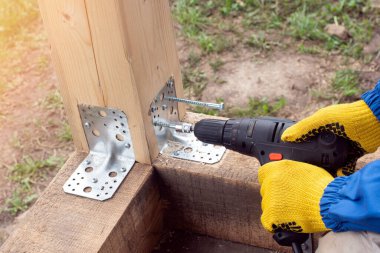 İşçi bir alet ve metal bir açı yardımıyla kereste çerçevesini güçlendirir. Sanatçı ahşap bir ev inşa ederken vidayı ahşap kirişle sıkıyor. .