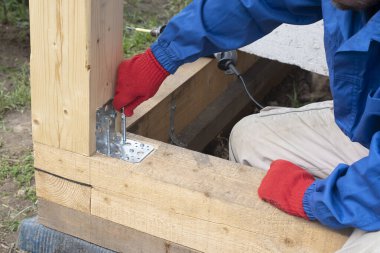 İşçi bir alet ve metal bir açı yardımıyla kereste çerçevesini güçlendirir. Sanatçı ahşap bir ev inşa ederken vidayı ahşap kirişle sıkıyor. .