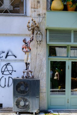 Klima ünitesinin üzerinde süslü bir avize tutan klasik bir erkek heykeli olan kumlu şehir manzarası eskimiş bir duvara karşı siyah grafiti ve açık yeşil bir mağaza vitriniyle çizilmiş..