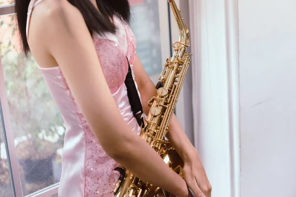 Young woman saxophonist with an instrument stands by the window. Cozy home interior. Concept of hobbies, inner world, development of talents, music school.