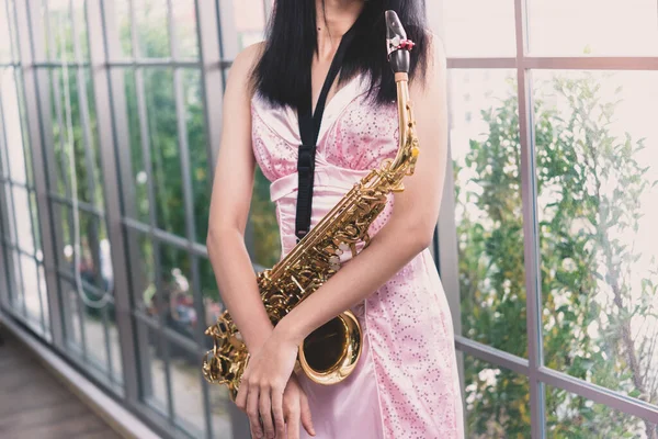Young woman saxophonist with an instrument stands by the window. Cozy home interior. Concept of hobbies, inner world, development of talents, music school.