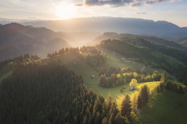 Gün batımında Transilvanya tepelerinin hava manzarası yeşil ormanlar ve dağ kenarındaki geleneksel kır evleri.