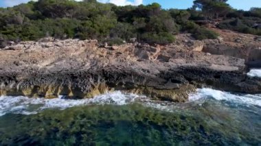 4K Hava Aracı 'nın Mallorca' da kristal berrak suyla çekilmiş videosu güneşli bir yaz gününde sert dalgalar kayalıklara çarpıyor.