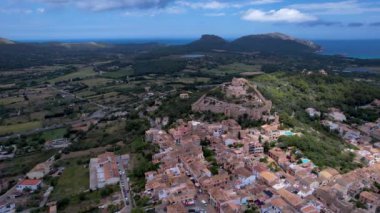 4K Hava Aracı, güneşli bir yaz gününde dağlarla ve denizlerle çevrili tepede devasa bir kalesi olan güzel bir terracotta renkli kasabasının videosu, Capdepera, Mallorca