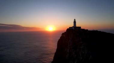 4K Aerial Drone 'un geniş deniz ufkundan yükselen ateşli kırmızı gündoğumu videosu kayalıkların kenarındaki deniz feneri Far of Cap de Formentor, Mallorca, İspanya