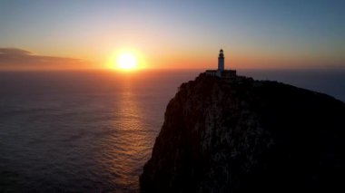 4K Aerial Drone 'un geniş deniz ufkundan yükselen ateşli kırmızı gündoğumu videosu kayalıkların kenarındaki deniz feneri Far of Cap de Formentor, Mallorca, İspanya