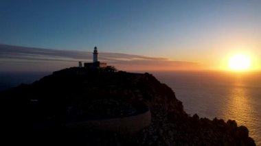 4K Aerial Drone 'un geniş deniz ufkundan yükselen ateşli kırmızı gündoğumu videosu kayalıkların kenarındaki deniz feneri Far of Cap de Formentor, Mallorca, İspanya