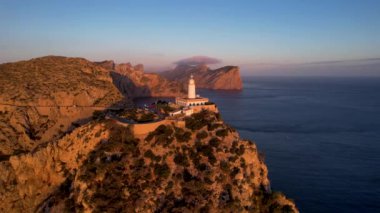 4K Aerial Drone 'un geniş deniz ufkundan yükselen ateşli kırmızı gündoğumu videosu kayalıkların kenarındaki deniz feneri Far of Cap de Formentor, Mallorca, İspanya