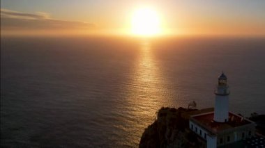 4K Aerial Drone 'un geniş deniz ufkundan yükselen ateşli kırmızı gündoğumu videosu kayalıkların kenarındaki deniz feneri Far of Cap de Formentor, Mallorca, İspanya