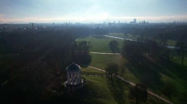 Monopteros ve Çin kulesinin 4K hava aracı videosu, Münih Almanya 'nın tarihi simgeleri güneşli ve sisli bir sonbahar gününde şehir panoramasına bakan İngiliz Bahçesi kamu parkı
