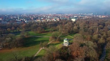 Monopteros ve Çin kulesinin 4K hava aracı videosu, Münih Almanya 'nın tarihi simgeleri güneşli ve sisli bir sonbahar gününde şehir panoramasına bakan İngiliz Bahçesi kamu parkı