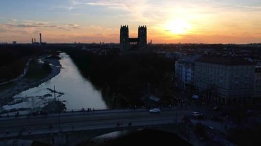 Münih Almanya 'nın Münih kentinde güneşli bir günde gün batımında şehir panoramasına bakan Isar nehrinin merkezindeki St. Maximilian Kilisesi' nin 4K hava aracı videosu.