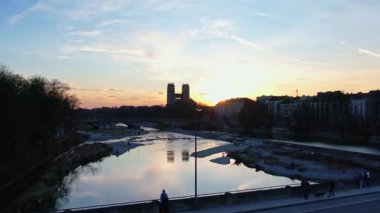 Münih Almanya 'nın Münih kentinde güneşli bir günde gün batımında şehir panoramasına bakan Isar nehrinin merkezindeki St. Maximilian Kilisesi' nin 4K hava aracı videosu.