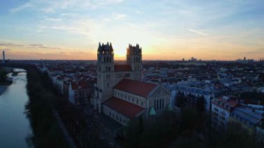 Münih Almanya 'nın Münih kentinde güneşli bir günde gün batımında şehir panoramasına bakan Isar nehrinin merkezindeki St. Maximilian Kilisesi' nin 4K hava aracı videosu.