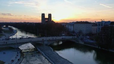 Münih Almanya 'nın Münih kentinde güneşli bir günde gün batımında şehir panoramasına bakan Isar nehrinin merkezindeki St. Maximilian Kilisesi' nin 4K hava aracı videosu.