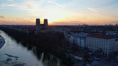 Münih Almanya 'nın Münih kentinde güneşli bir günde gün batımında şehir panoramasına bakan Isar nehrinin merkezindeki St. Maximilian Kilisesi' nin 4K hava aracı videosu.
