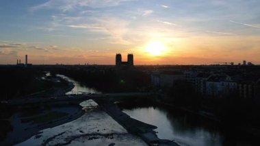 Münih Almanya 'nın Münih kentinde güneşli bir günde gün batımında şehir panoramasına bakan Isar nehrinin merkezindeki St. Maximilian Kilisesi' nin 4K hava aracı videosu.