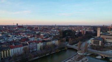 Münih Almanya 'nın Münih kentinde güneşli bir günde gün batımında şehir panoramasına bakan Isar nehrinin merkezindeki St. Maximilian Kilisesi' nin 4K hava aracı videosu.