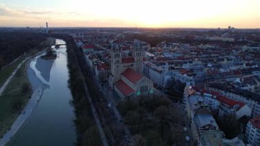 Münih Almanya 'nın Münih kentinde güneşli bir günde gün batımında şehir panoramasına bakan Isar nehrinin merkezindeki St. Maximilian Kilisesi' nin 4K hava aracı videosu.
