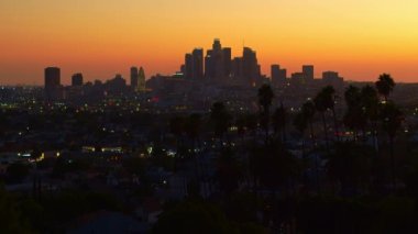Los Angeles şehir merkezinin 4K görüntüsü. Parlayan ışıklar ve reklamlarla gökdelenlerin üzerinde güzel bir günbatımı. Ön planda palmiye ağaçları, renkli bir metropol gökyüzünün önünde. Kaliforniya, ABD