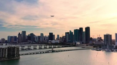 4K hava görüntüsü, körfez manzarası ve Miami şehir merkezinin modern mimarisine sahip binalar. Uçak şehrin üzerinde pembe bulutlu mavi gökyüzünün arka planına doğru uçuyor. Renkli manzara