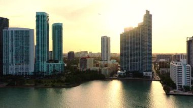 4K hava görüntüsü, Miami 'deki sahil şeridi manzarası, yüksek yerleşim yerleriyle modern binalar. Günbatımının altın ışıkları çerçevedeki bulutların arasından parlıyor. Arka planda bir uçak uçuyor.