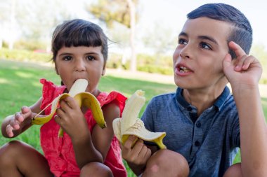 Two kids eating banana sitting outdoors on the grass in a park. Childhood and food concept.