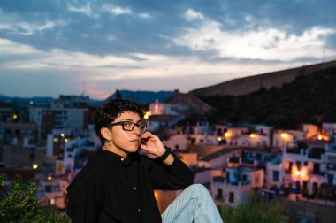 Genç bir transseksüel gün batımında arka planda bir köyün görüntüsüyle dışarıda poz verirken yukarı bakıyor..